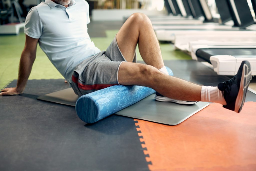 Unrecognizable mature sportsman using foam roller for stretching leg muscles during gym workout.